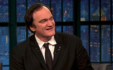 De terno, Quentin Tarantino sorri sem mostrar os dentes em um cenário de talk show