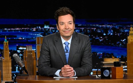 Jimmy Fallon força sorriso, com expressão contrariada, no cenário do The Tonight Show