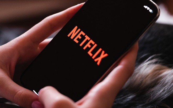 Imagem de uma mão mexendo no celular com o logo da Netflix, serviço de streaming, na tela
