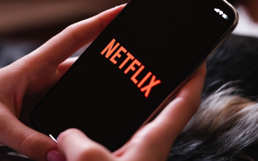Imagem de uma mão mexendo no celular com o logo da Netflix, serviço de streaming, na tela