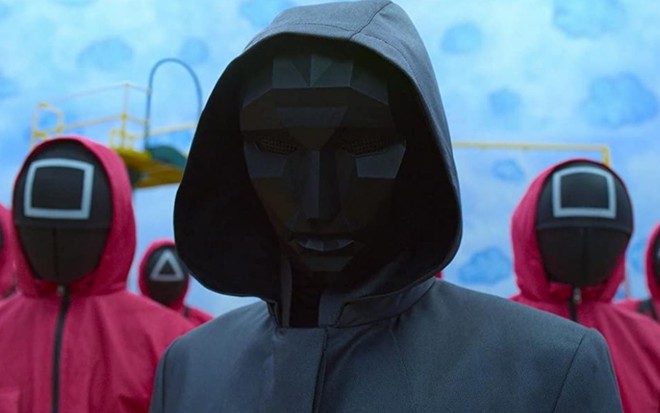 Mascarado de Round 6, da Netflix, usa uma roupa toda preta e está acompanhado de pessoas mascaradas com trajes vermelhos