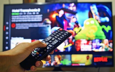 Mão segunda controle remoto diante de uma smart TV que mostra um catálogo de streaming