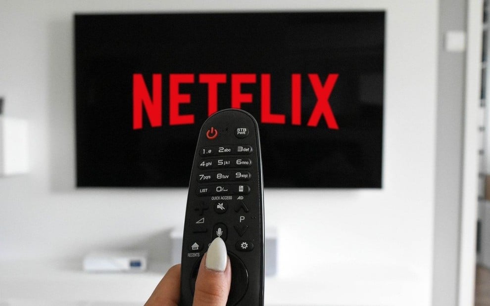 Uma mão segura um controle remoto apontado para uma televisão que tem o logotipo da Netflix