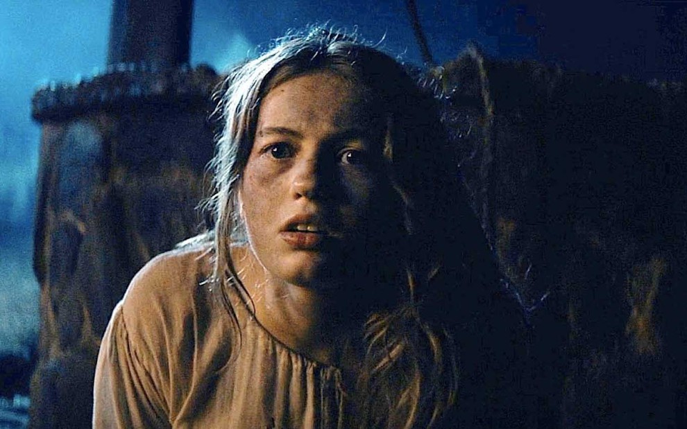A atriz Kiana Madeira como Deena está com uma tocha na mão direita e com uma veste parda sobre o corpo dentro de uma caverna em cena de Rua do Medo