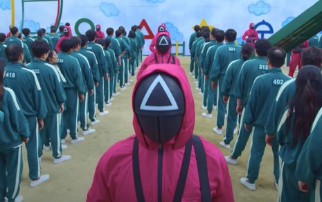 Cena de Round 6, série da Netflix: pessoas encapuzadas de vermelho e com uma máscara preta; ao lado delas, dezenas de pessoas enfileiradas