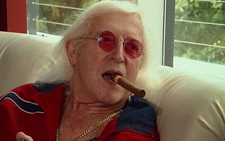 Jimmy Savile (1926-2011) de óculos vermelhos e charuto na boca sentado em um sofá branco