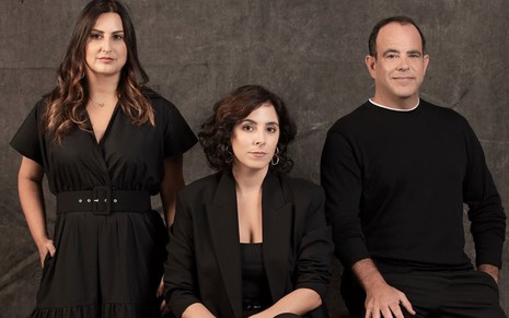 A jornalista Daniela Arbex, a diretora Julia Rezende e o roteirista Gustavo Lipsztein, um ao lado do outro, todos sérios e de roupas pretas, em foto de divulgação com fundo cinza