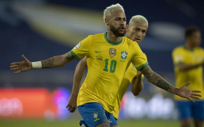 Neymar com a camisa amarela e o short azul da seleção brasileira. Ele comemora um gol e é observado por Richarlison.