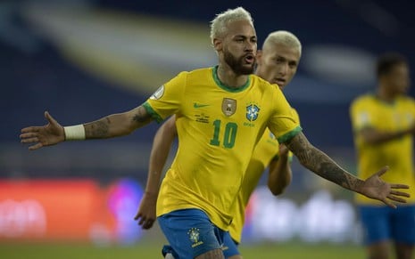 Neymar com a camisa amarela e o short azul da seleção brasileira. Ele comemora um gol e é observado por Richarlison.
