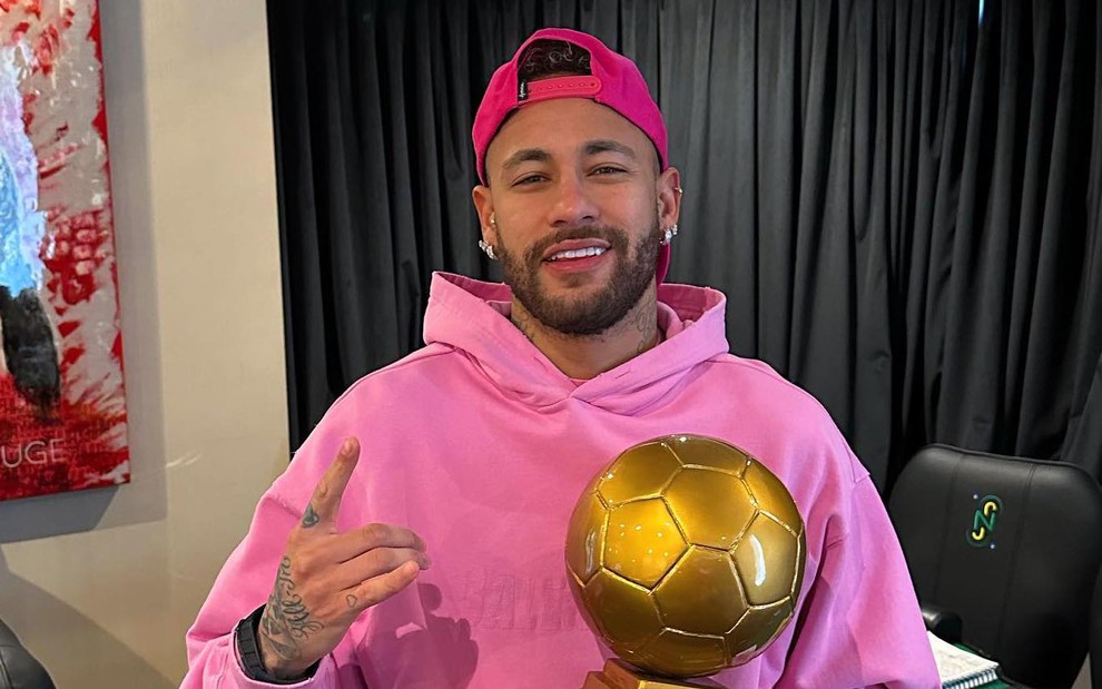 O jogador Neymar usa agasalho rosa, segura uma taca em formato de bola e está também com boné pink