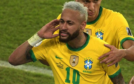 Neymar com a camisa amarela e o short azul da seleção brasileira. Ele comemora um gol e é observado por Richarlison.