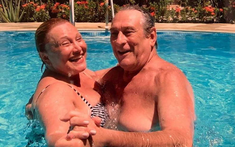 Os atores Nicette Bruno e Paulo Goulart; eles estavam abraçados e de mãos dadas, dentro de uma piscina, olhando para a foto e sorrindo