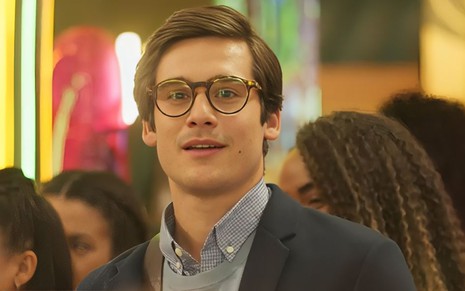 Em cena de Fuzuê, Nicolas Prattes usa óculos, blusa, colete de tricô e blazer; ele está parado, sorrindo, olhando para alguém
