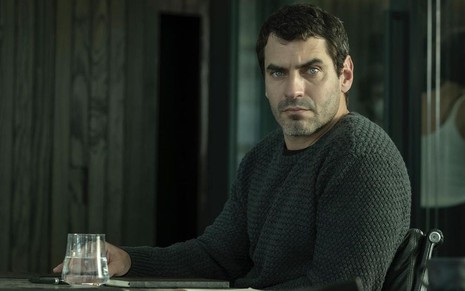 Nikolas Antunes está com tricô verde escuro, sentado em uma cadeira e há um copo de vidro na mesa em que ele apoia o braço