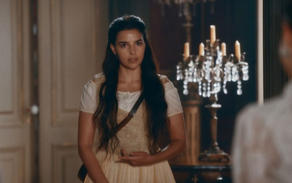 Pilar (Gabriela Medvedovski) está em pé na sala de Luísa (Mariana Ximenes), há um castiçal atrás da jovem em cena de Nos Tempos do Imperador