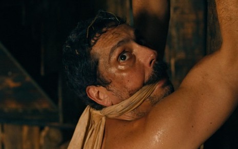Tonico (Alexandre Nero) está sujo e suado, com mordaça e as mãos amarradas em cena de sequestro em celeiro em Nos Tempos do Imperador