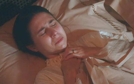 Atriz Daphne Bozaski aparece deitada em cama, com os olhos fechados e uma expressão de pânico em cena de Nos Tempos do Imperador