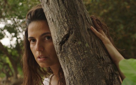 Emanuelle Araujo tem visual de mulher brejeira, com expressão assustada e meio escondida atrás de uma árvore