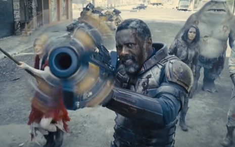 Idris Elba usando uma arma em cena de O Esquadrão Suicida; ele está acompanhado de outros personagens