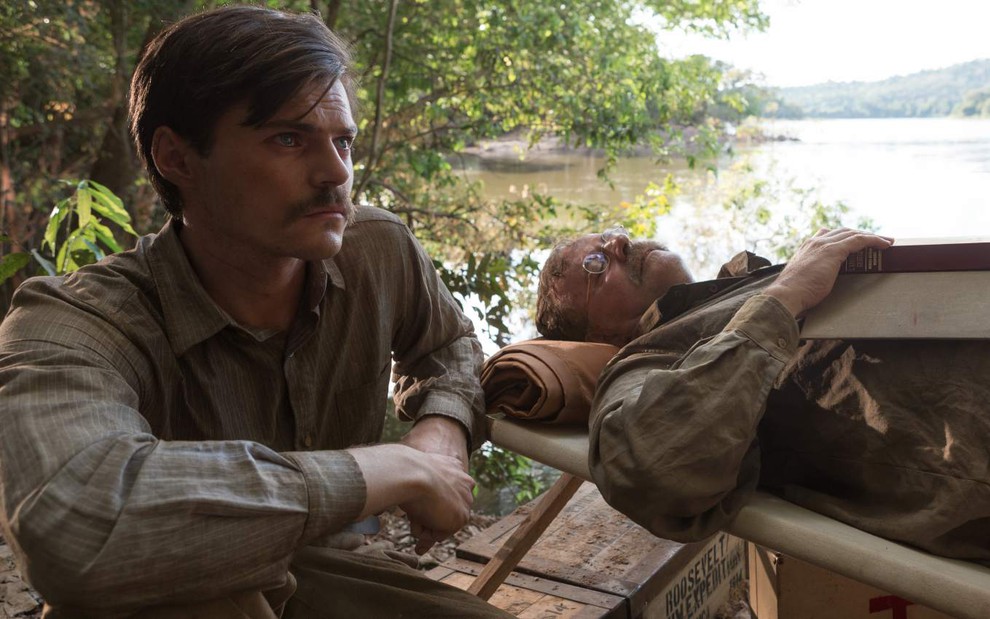 Os atores estrangeiros Chris Mason e Aidan Quinn lado a lado, Mason sentado e Quinn deitado, em região amazônica, com floresta e rio no plano de fundo