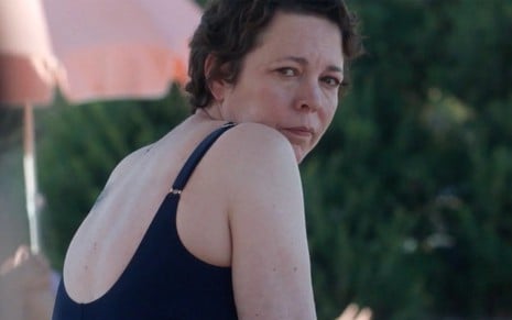 Olivia Colman de maiô preto, expressão séria, olha para trás em cena como a protagonista Leda do filme A Filha Perdida, estreia da Netflix