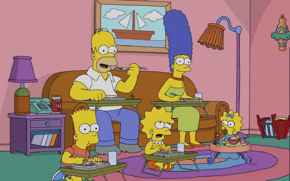 A família Simpson na sala. Homer come algo, Margie sorri para a câmera, enquanto Bart, Lisa e Maggie estão brincando no chão da sala da casa do desenho animado.