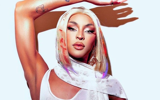 Foto mostra Pabllo Vittar vestindo regata branca; ela está parada na frente de uma parede branca