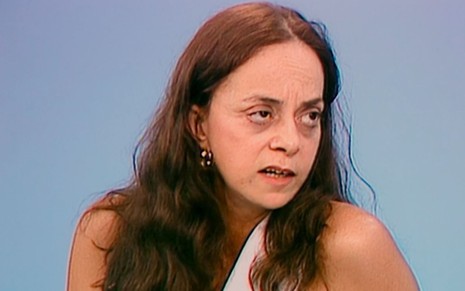 A autora Gloria Perez nos anos 1990, de cabelos compridos, expressão séria, em frente a fundo azul