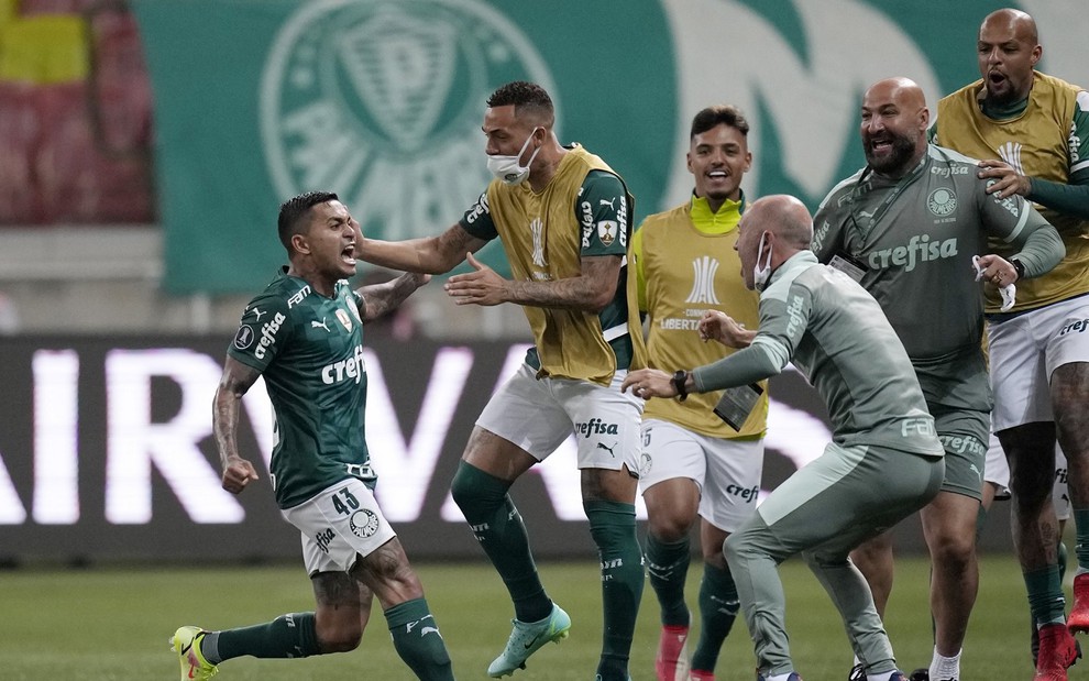 Dudu vibra com gol do Palmeiras. Ele usa uma camisa verde e comemora com seus colegas de banco, que usam um colete amarelo