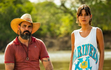 Os atores Juliano Cazarré e Julia Dalavia, caracterizados como seus personagens em Pantanal, estão à beira de um rio. Ele usa chapéu, e ela veste uma camiseta moderna com top aparecendo