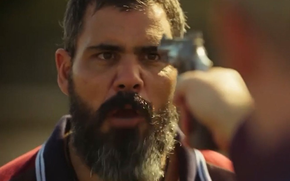 O ator Juliano Cazarré como Alcides em Pantanal; ele está olhando para a frente, com a boca semiaberta enquanto conversa e olha para uma arma que está sendo apontada em sua direção