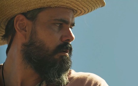 O ator Juliano Cazarré como Alcides em Pantanal; ele está de chapéu, olhando para o lado, com cara séria e pensativa