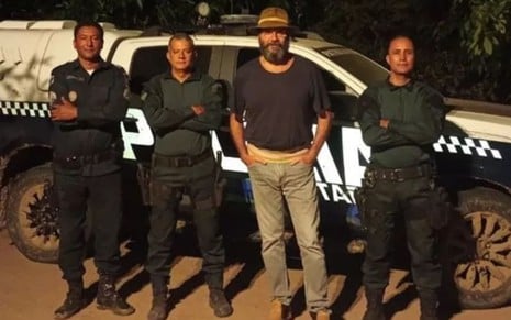 Três policiais militares e Almir Sater posam em frente ao carro da PM; ator tem a barba longa e veste camisa preta, calça bege e botinas. Os três profissionais estão uniformizados e posam de braços cruzados.