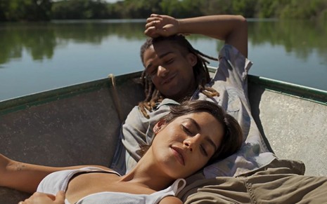 Os atores Julia Dalavia e Lucas Leto como Guta e Marcelo em Pantanal; na foto eles estão de olhos fechados, deitados em um barco, com ela deitada sobre suas pernas enquanto ele sorri