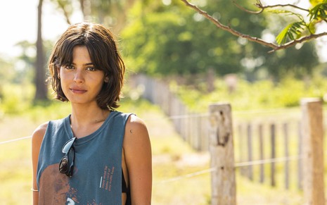 A atriz Julia Dalavia está caracterizada como sua personagem em Pantanal com uma camiseta cavada e óculos pendurado