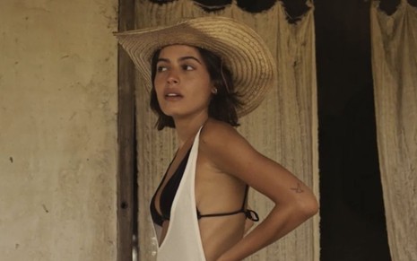 A atriz Julia Dalavia está caracterizada como sua personagem em Pantanal com chapéu, biquíni e uma camiseta cavada por cima