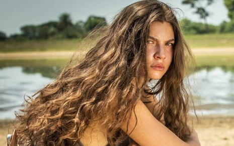 A atriz Alanis Guillen com cabelos compridos e soltos, olhar sério, posa para câmera em foto de divulgação de Pantanal, com paisagem ao fundo