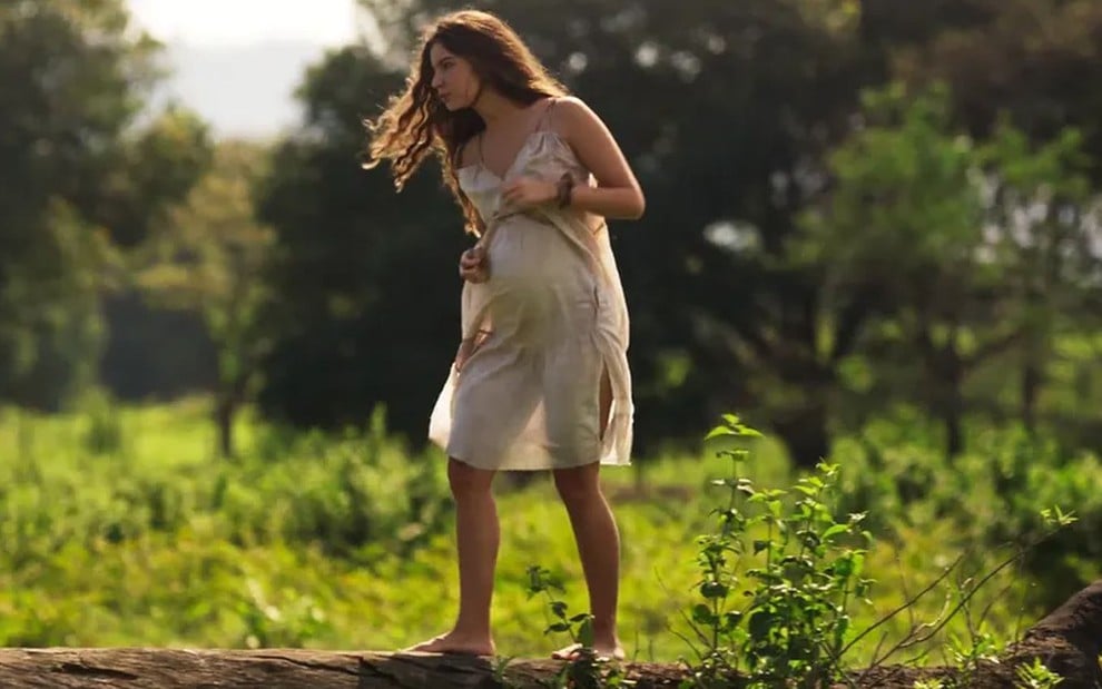 A atriz Alanis Guillen como Juma em Pantanal; ela está de lado, andando em cima de um tronco enquanto olha para frente e segura a barriga
