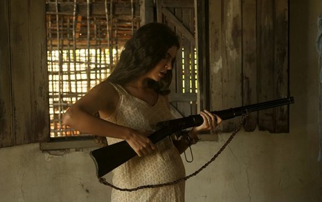 A atriz Alanis Guillen como Juma em Pantanal; ela está de lado, segurando uma espingarda e olhando para a arma enquanto a prepara