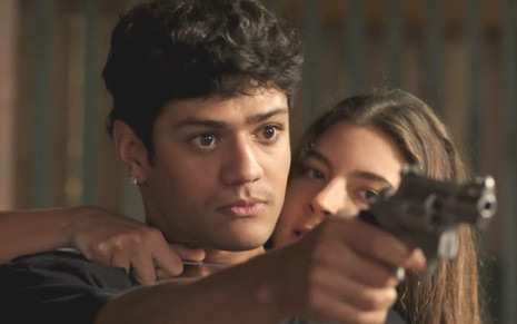 Os atores Gabriel Santana e Alanis Guillen como Renato e Juma em Pantanal; ele está apontando uma arma para frente enquanto ela está por trás com uma faca em seu pescoço