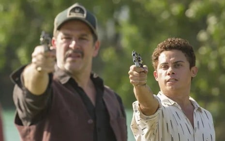 Tenório (Murilo Benício) está com arma apontada para frente, e Zaquieu (Silvero Pereira) aparece atrás com revólver na mão em cena da novela Pantanal