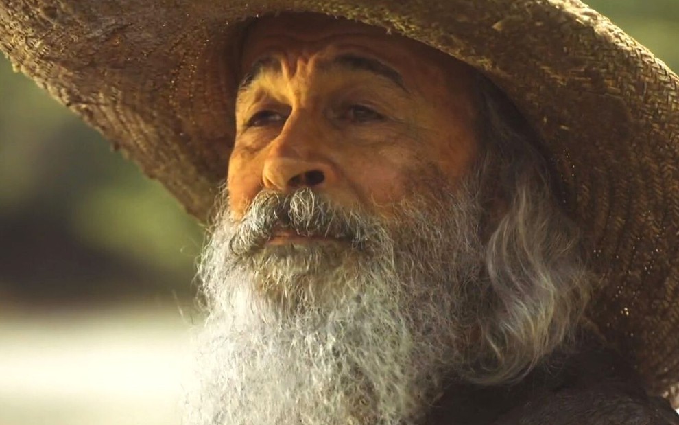 Osmar Prado usa chapéu, barba comprida e segura um galho na mão; ele está caracterizado como Velho do Rio em cena de Pantanal, novela das nove da Globo