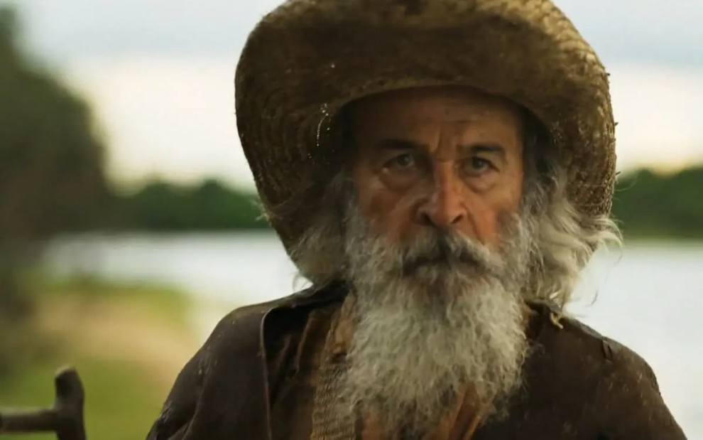 O ator Osmar Prado de chapéu, barba branca e expressão carrancuda, na beira de rio, em cena de Pantanal