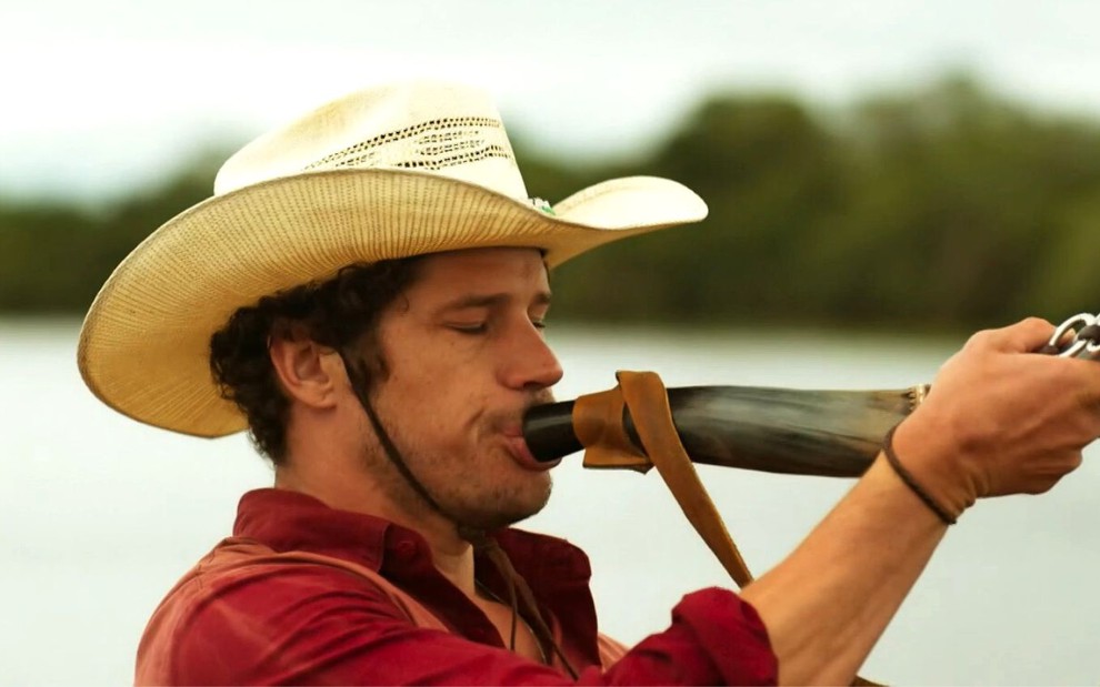 O ator José Loreto como Tadeu em Pantanal; ele está de chapéu e de lado, tocando berrante