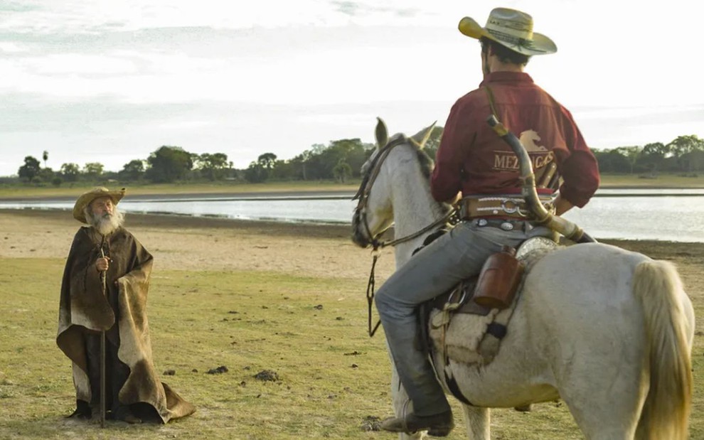 Os atores Osmar Prado e José Loreto como Velho do Rio e Tadeu em Pantanal; eles estão frente a frente, enquanto o primeiro está parado, segurando um cajado, e o segundo está de costas, montado em um cavalo, olhando para ele