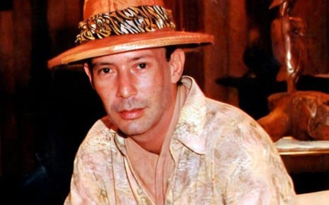 O ator João Alberto Pinheiro caracterizado como Zaquieu, com chapéu, lenço com estampa animal e camisa bege clara estampada, em gravação de Pantanal