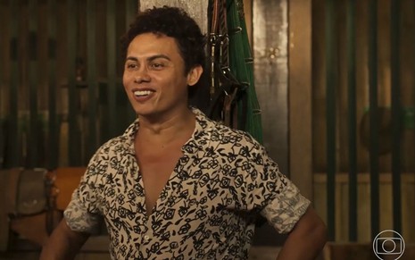 O ator Silvero Pereira como Zaquieu em Pantanal; ele está olhando para frente e sorrindo enquanto conversa no galpão