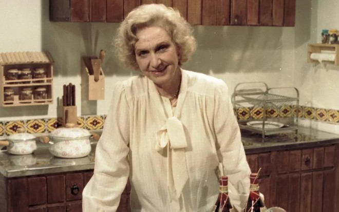 Lélia Abramo (1911-2004) como a personagem Vitória da novela Pão Pão, Beijo Beijo (1983), com camisa branca com laço, em cenário de cozinha