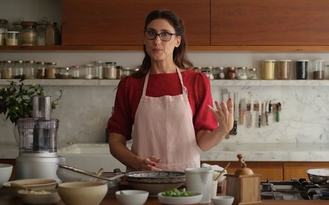 A cozinheira Paola Carosella em uma cozinha, com cabelos presos, óculos de grau, blusa vermelha e um avental rosa