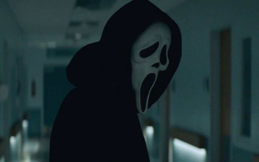 Imagem do personagem Ghostface, antagonista do novo filme de Pânico; de pé em um corredor escuro
